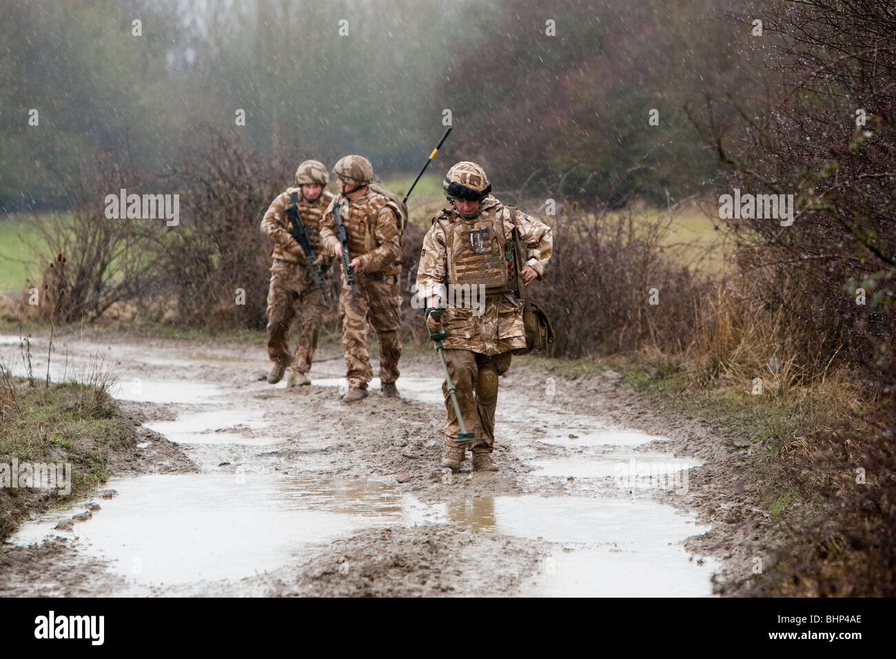 Three British soldier walking in line along a muddy road behind a metal detector looking for an IED roadside bomb Stock Photo