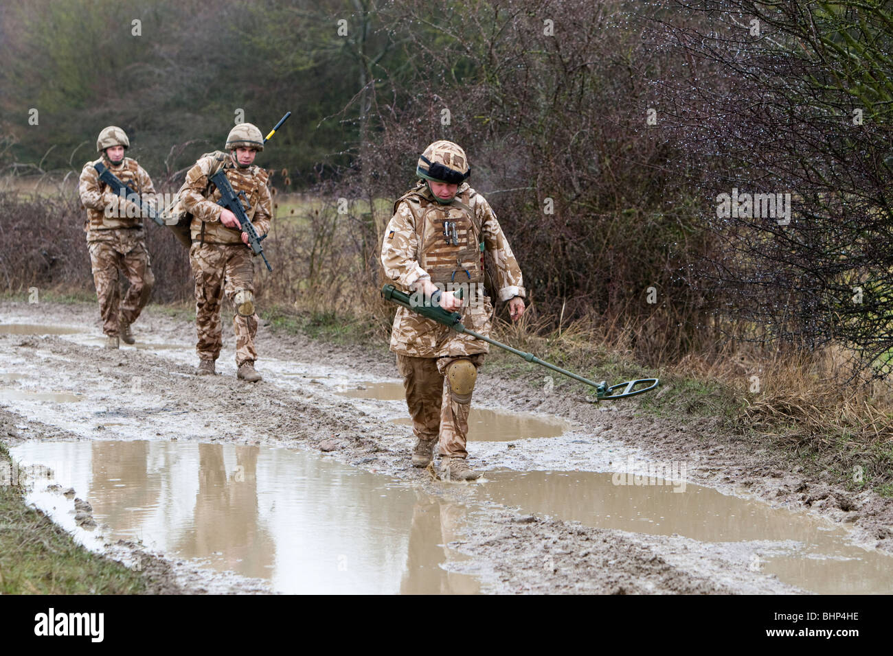 Three British soldier walking in line along a muddy road behind a metal detector looking for an IED roadside bomb Stock Photo