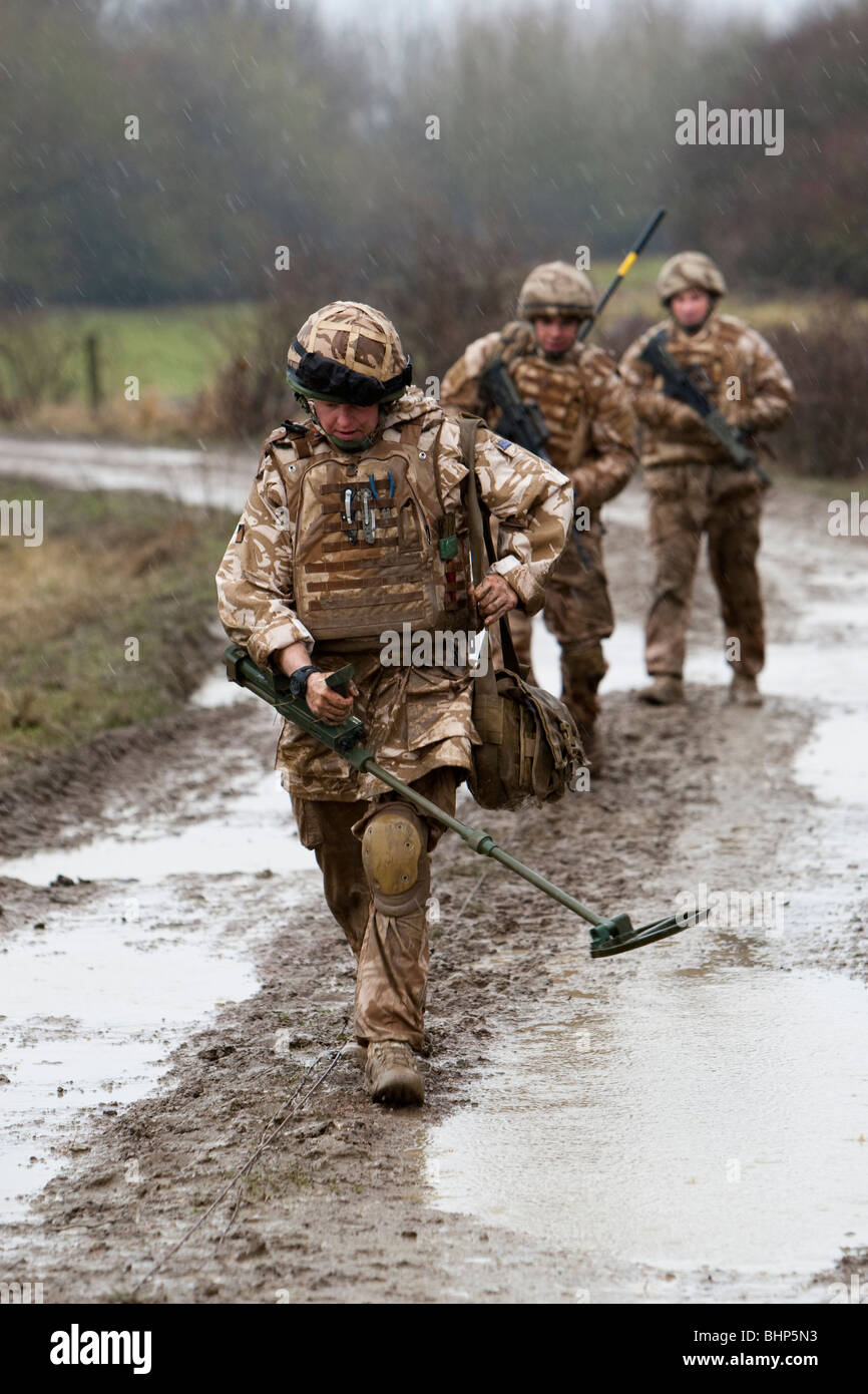 Three British soldier walking in line along a muddy road behind a metal detector looking for an IED roadside bomb Stock Photo