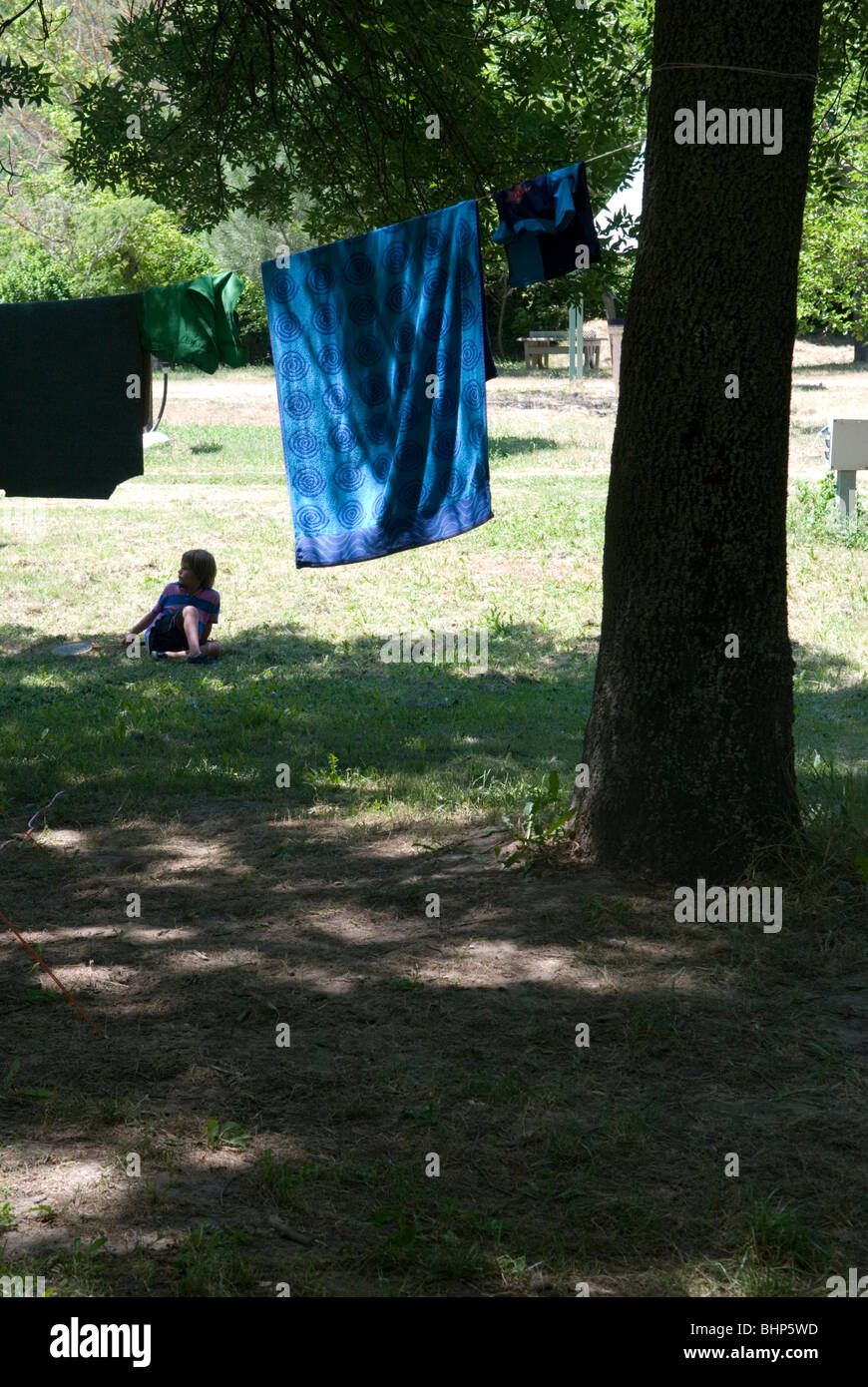 7 year old young boy with frisbee sits on grass in the shade beneath a washing line with drying towels on a campsite in france Stock Photo