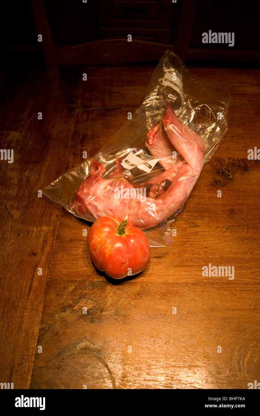 a slighty macabre still life of a large steak tomato and a skinned rabbit in a bag on a wooden kitchen table Stock Photo