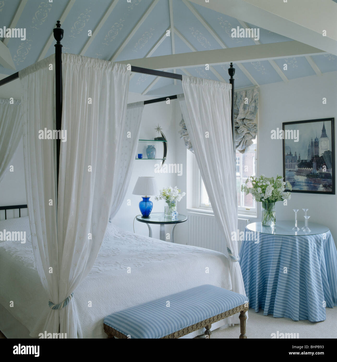 Blue upholstered stool below simple four-poster bed with white drapes and linen in white bedroom with blue cloth on round table Stock Photo