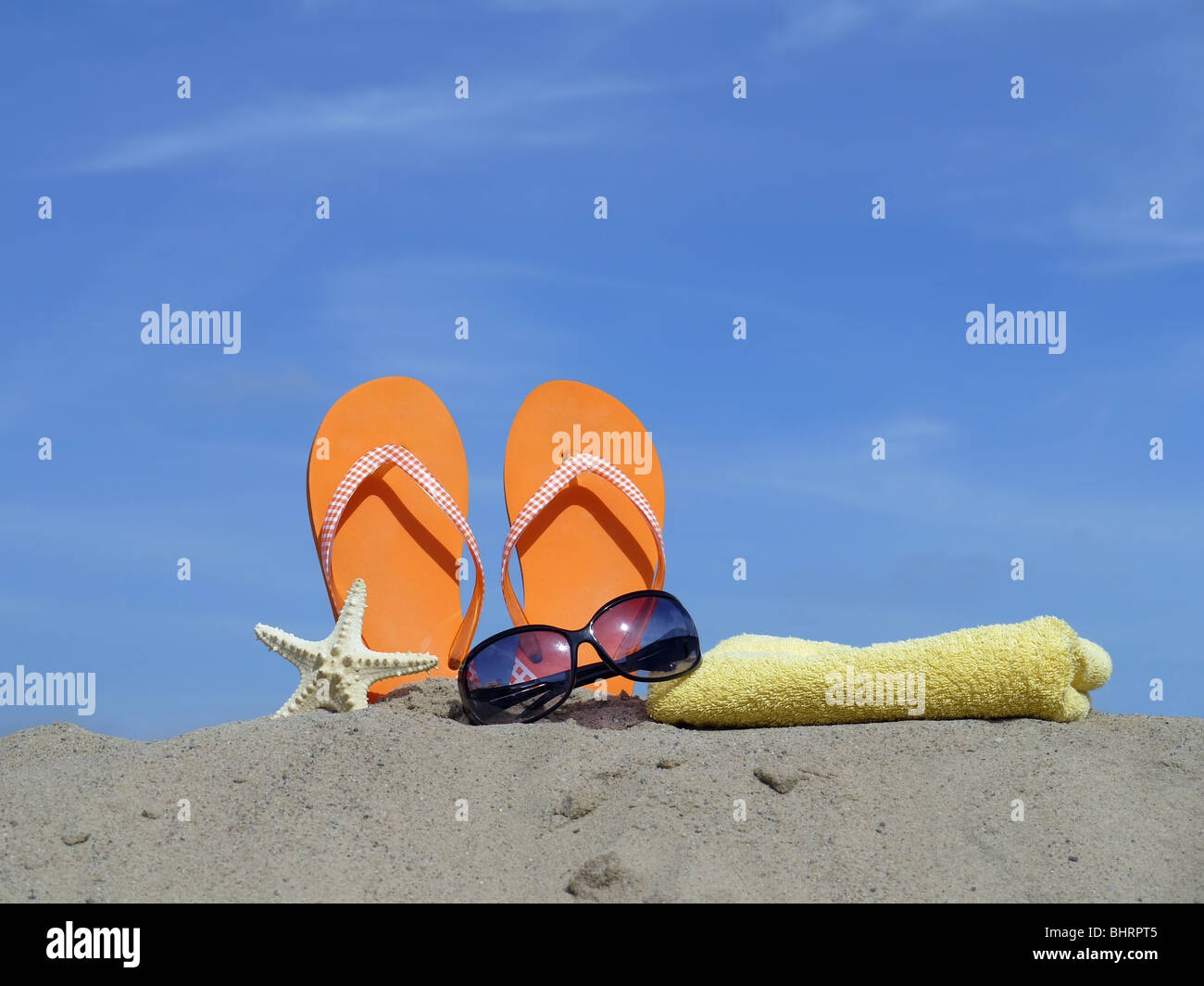 Orange flip-flops driven vertically into beach sand, starfish, sunglasses and yellow bath towel over blue sky Stock Photo