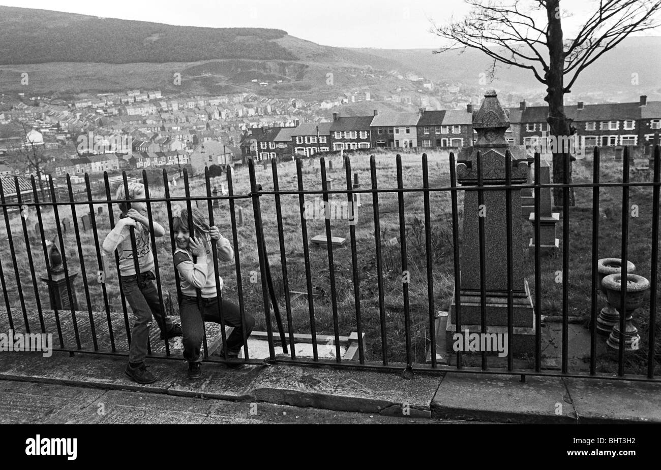 Two local children squeeze through iron railings of an unkempt Welsh cemetery. Stock Photo