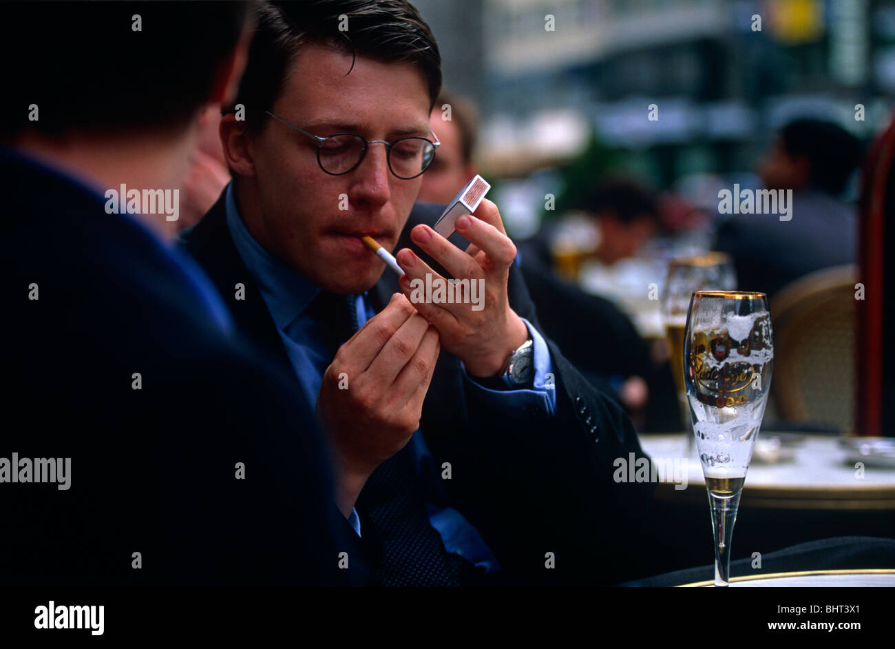 Striking a light in an outdoor café, a young businessman puts a match to his cigarette as a colleague talks in Frankfurt. Stock Photo