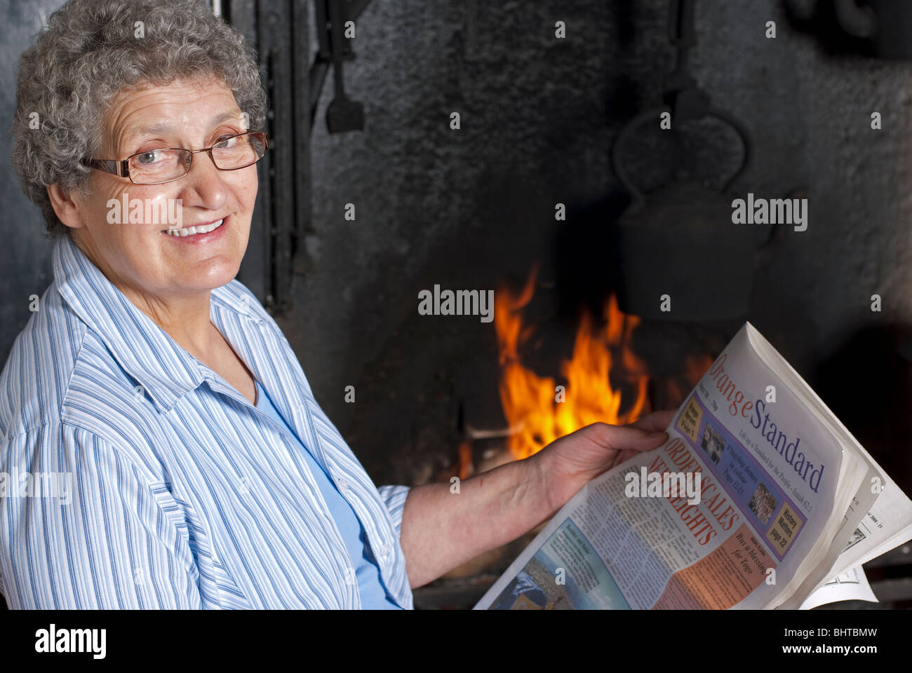Woman seated by the cottage fireside reading a newspaper with a glint in her eye Stock Photo