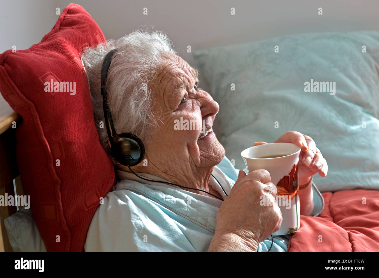 ELDERLY LADY CARE CARER Happy content elderly lady resting in bed wearing headphones enjoys a cup of tea in her sunny comfortable room Stock Photo