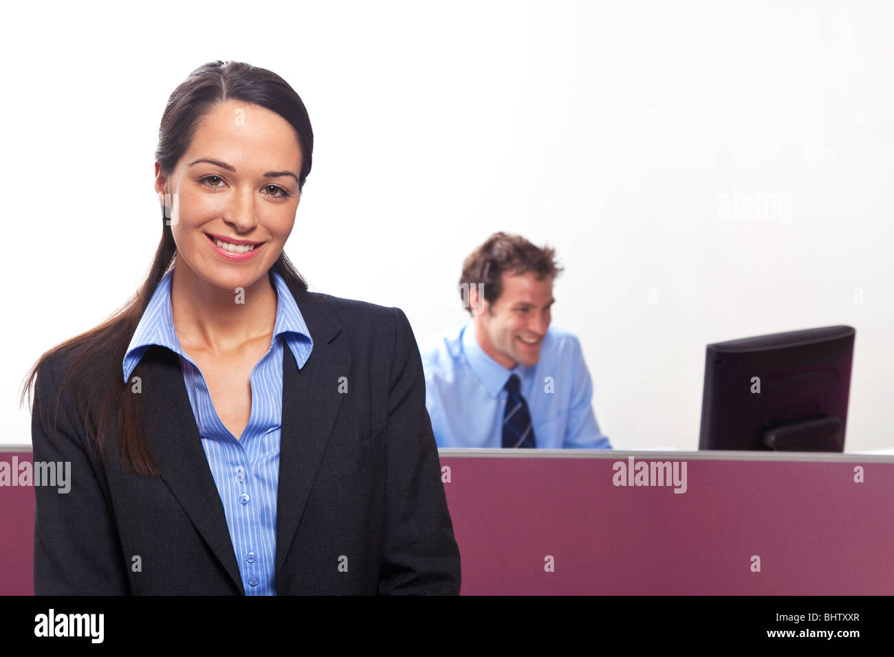 A businesswoman smiling towards camera, her colleague sat at a desk behind. Stock Photo