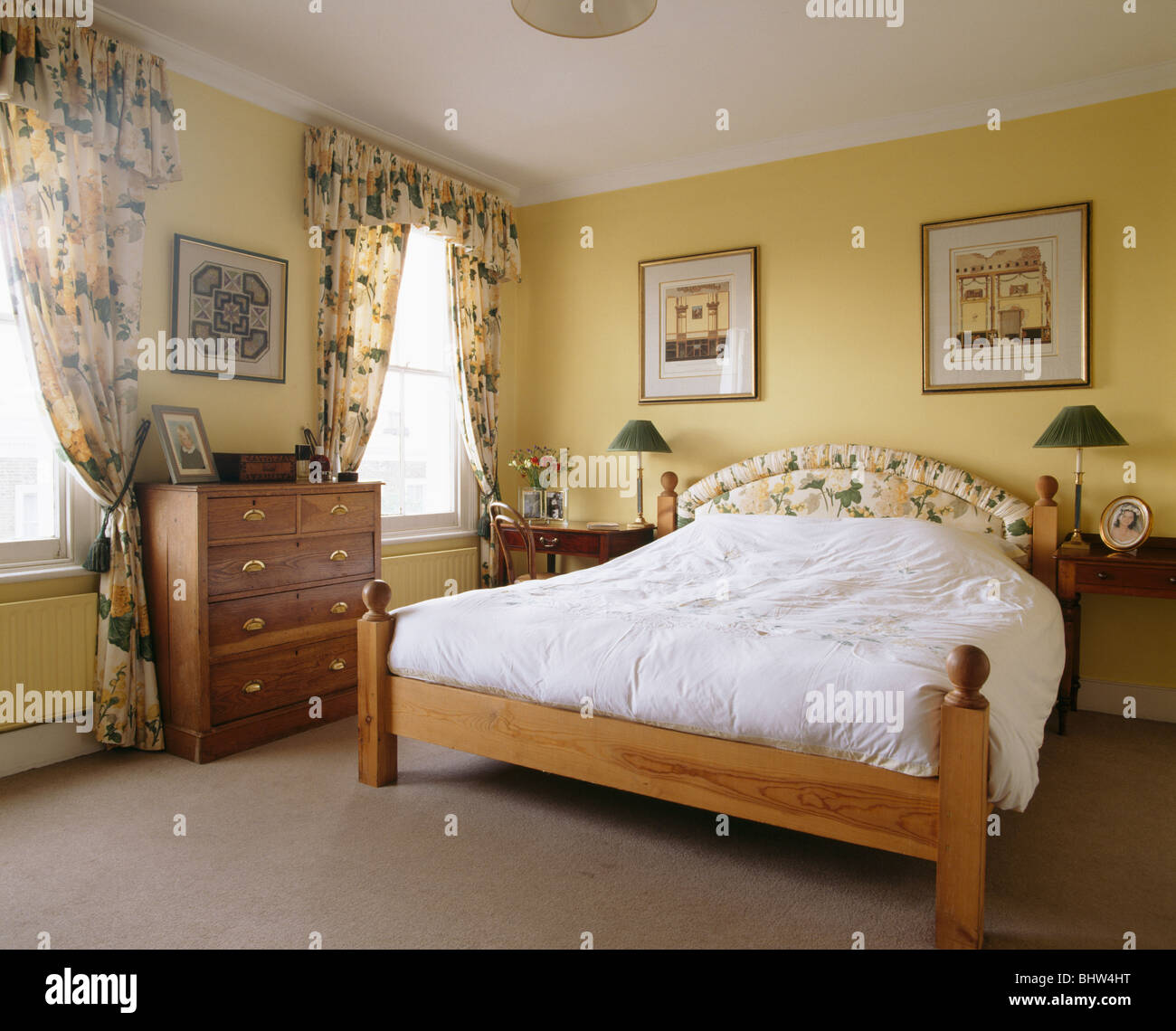 Pictures on wall above pine bed with white bedlinen in traditional yellow  bedroom with yellow floral curtains Stock Photo - Alamy