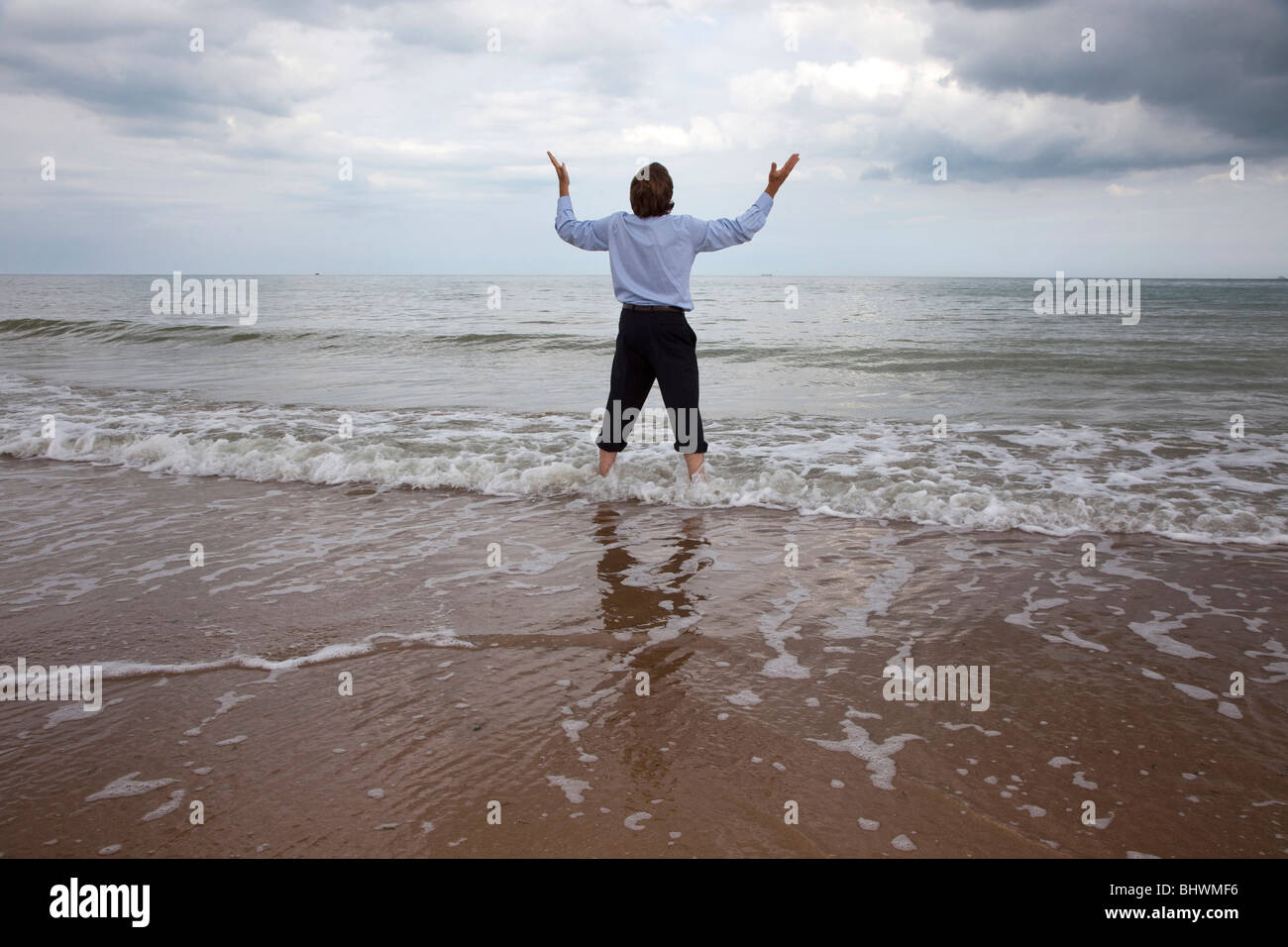 Back view of a man standing in the sea, arms up looking to the sky in despair Stock Photo