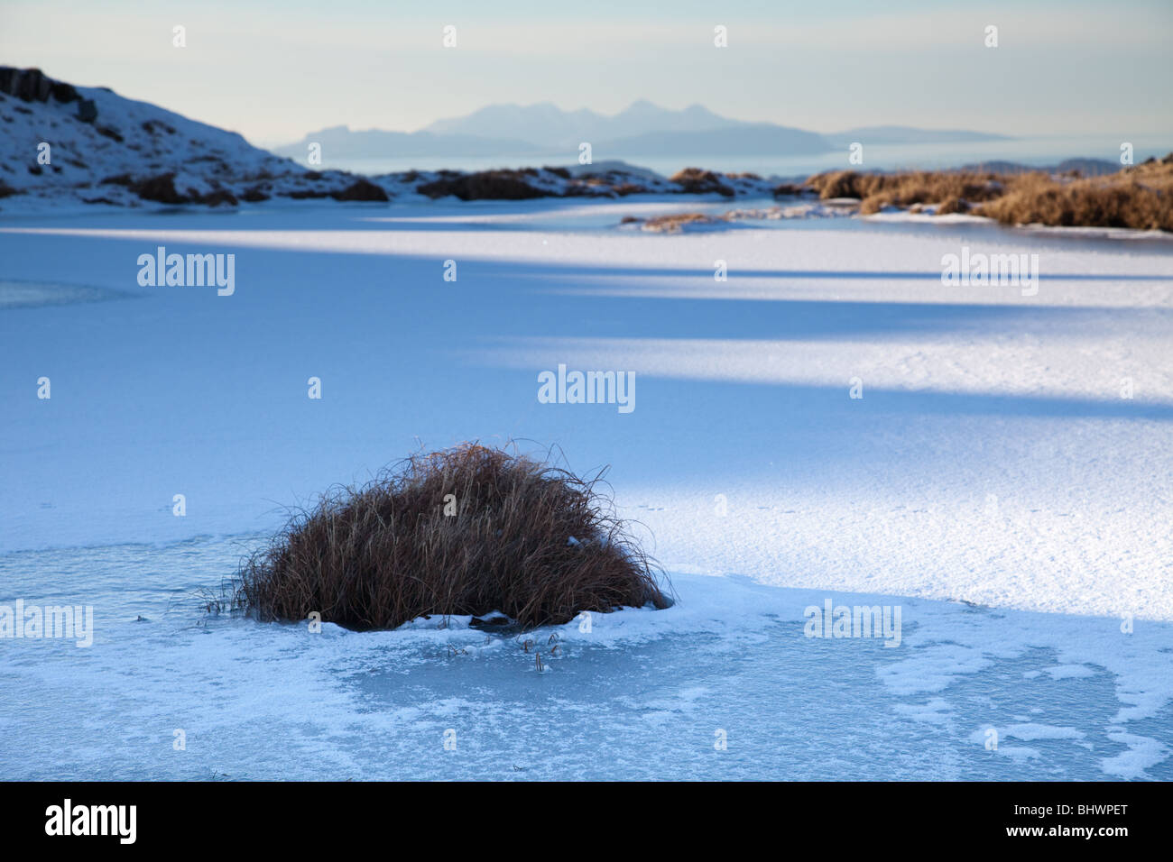 The Cuillin hills of Rum seen in the distance over a frozen loch during the descent of Beinn Resipol Stock Photo