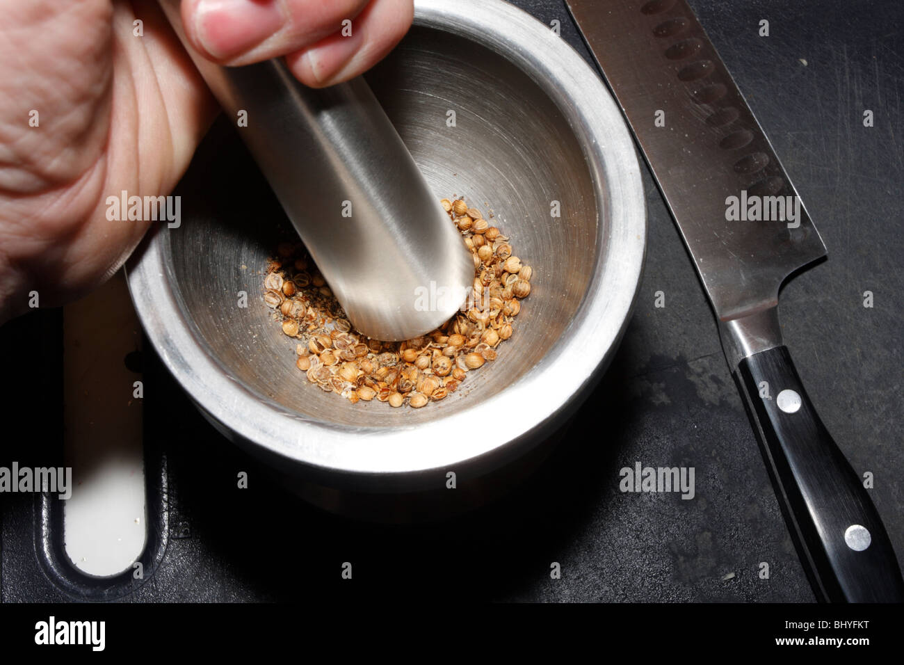 Hand crushing coriander seeds in mortar with pestle, stainless steel, next to chopping knife on black plastic cutting board. Stock Photo