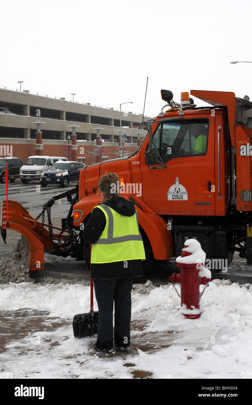 Woman with snow shovel watches from sidewalk as municipal truck with snowplow blade turns a corner. Stock Photo