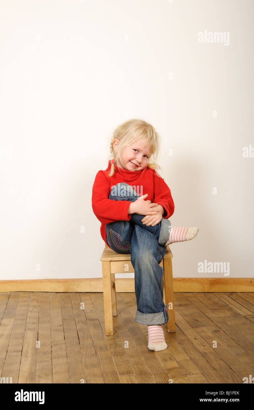 Stock photo of a four year old girl sitting on a wooden chair with her legs crossed. She has a cheeky expression on her face. Stock Photo