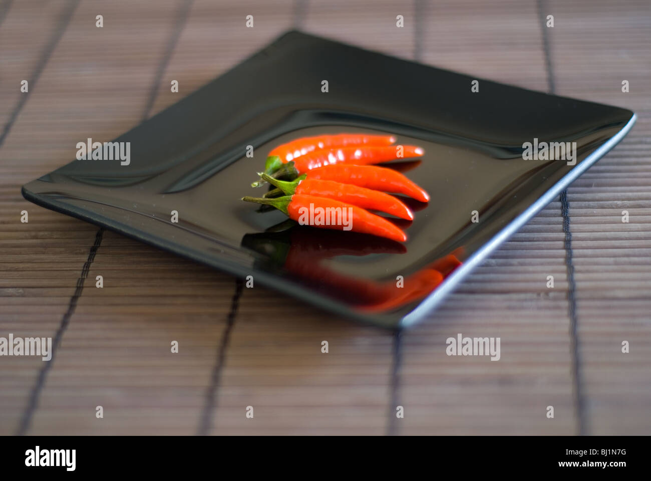 An Asian Style Square Black Plate with Four Red Chillis on a bamboo surface Stock Photo