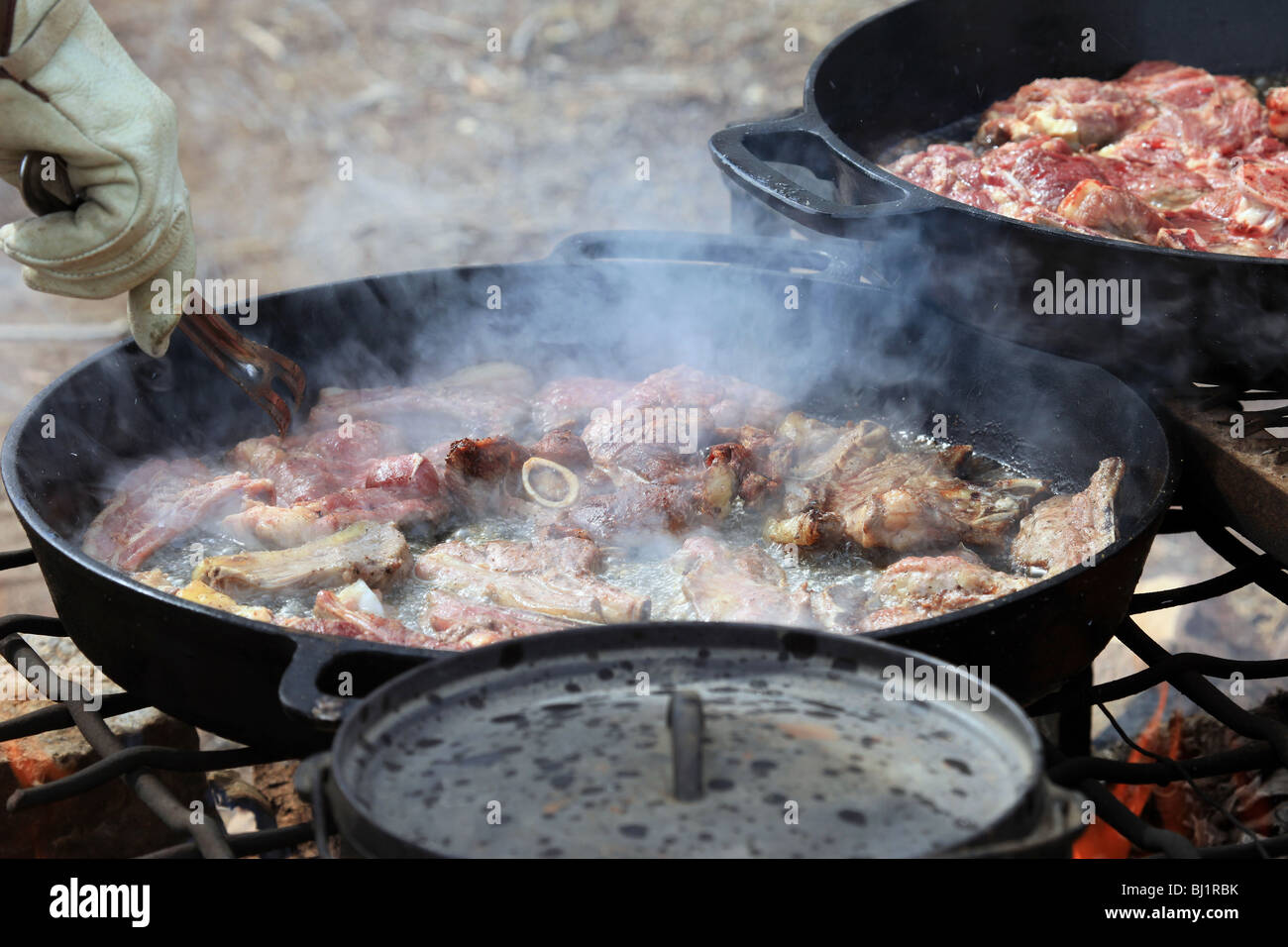 Mutton and lamb steaks and chops cooking in cast iron pans over an open