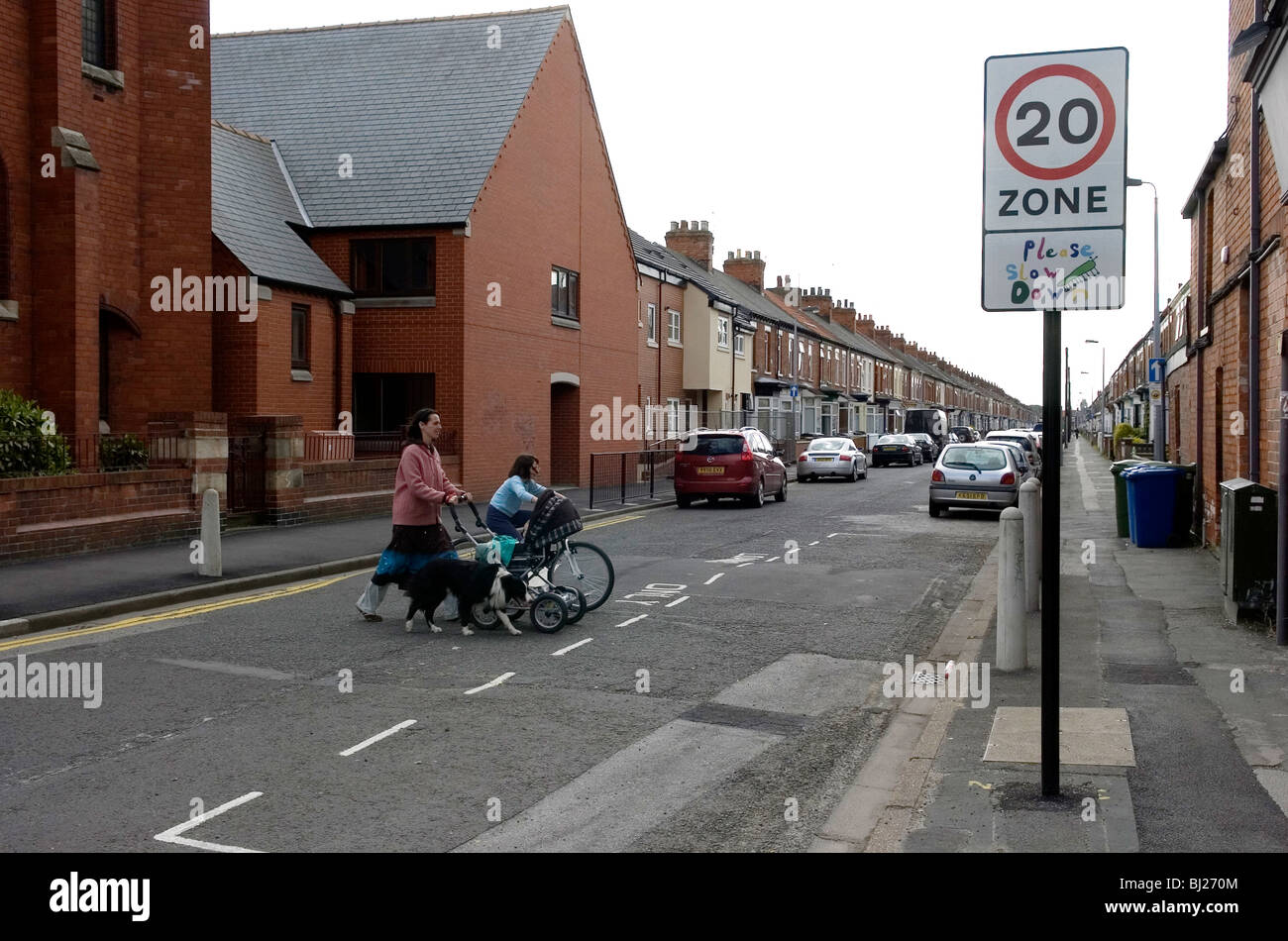 A woman pushes a pram across a street in Hull, UK, with a young girl on a bicycle. The street has a 20 mile per hour sign. Stock Photo