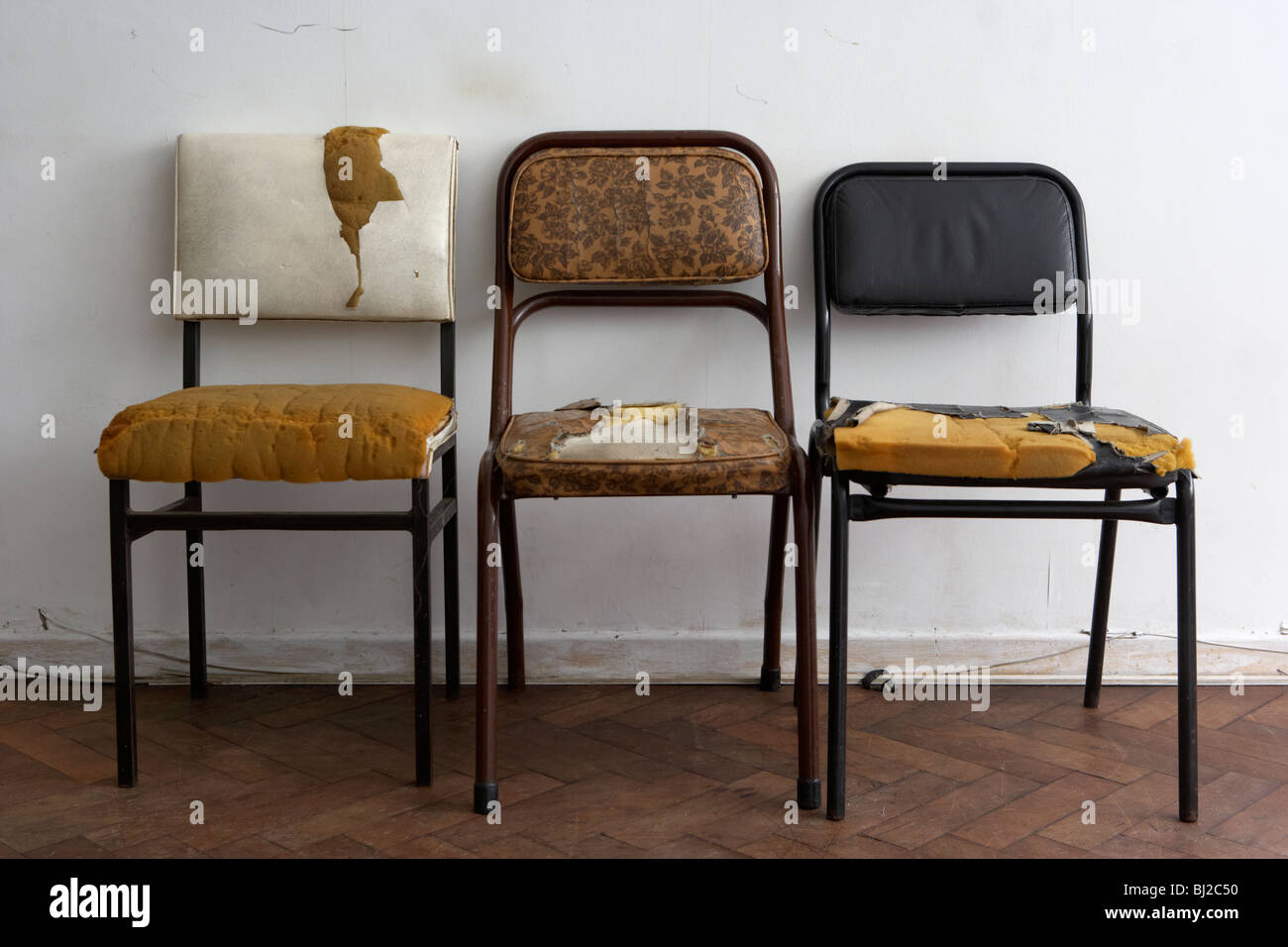 three old dirty worn out chairs against a white wall in a run down