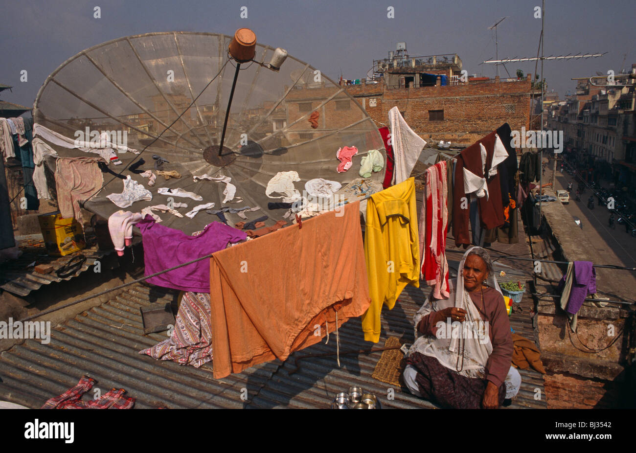 A Nepali lady sits on corrugated iron alongside a giant satellite dish on the roof of her home in a suburb of Kathmandu. Stock Photo