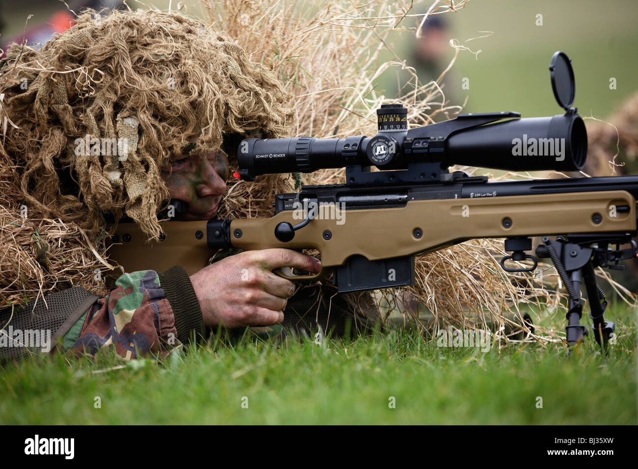 Lying on his stomach, a camouflaged British infantry sniper is seen looking down the telescopic sight of a L115A3 rifle. Stock Photo