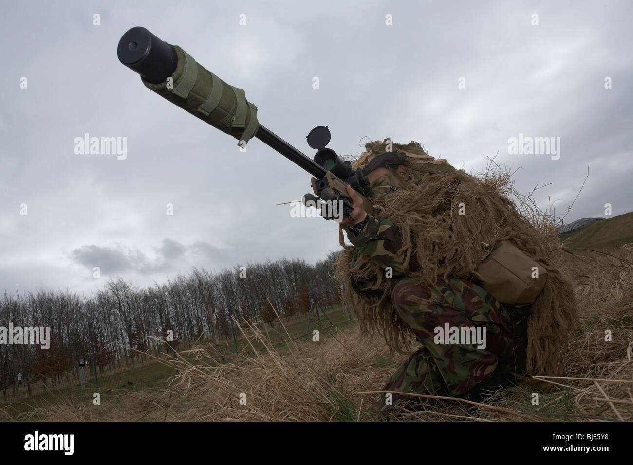 A camouflaged British infantry soldier is seen looking down the telescopic sight of a L115A3 sniper rifle. Stock Photo