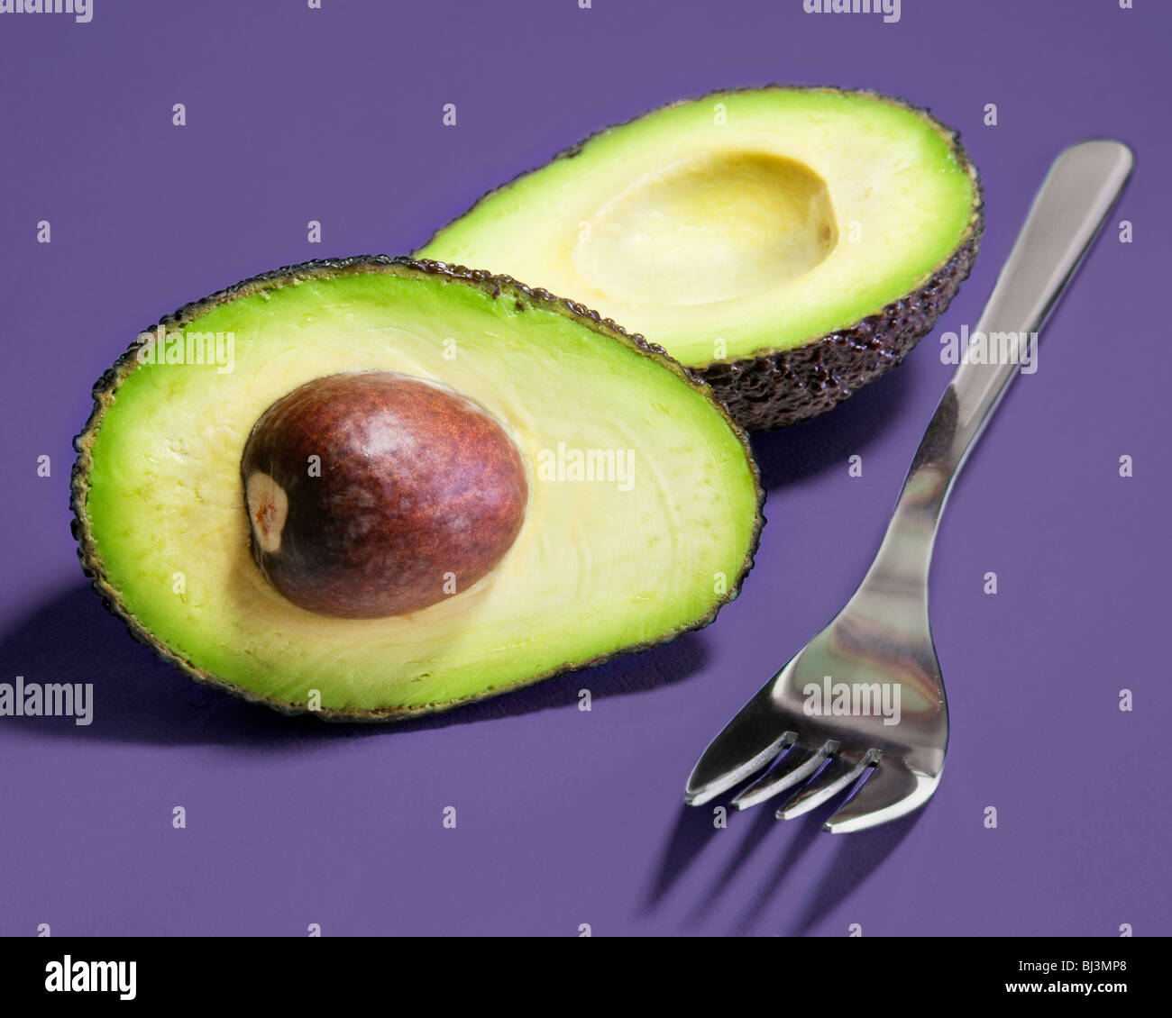 Hass variety avocado pear sliced into two halves along with a fork against a lilac background. Stock Photo