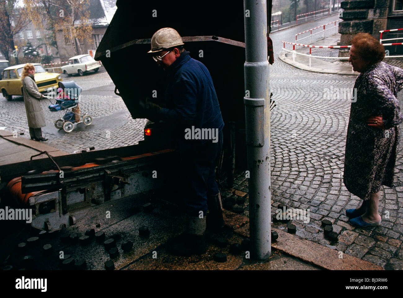 A coal delivery man deposits chunks of brown coal into the cellar via a conveyor belt for an elderly lady who stands outside. Stock Photo