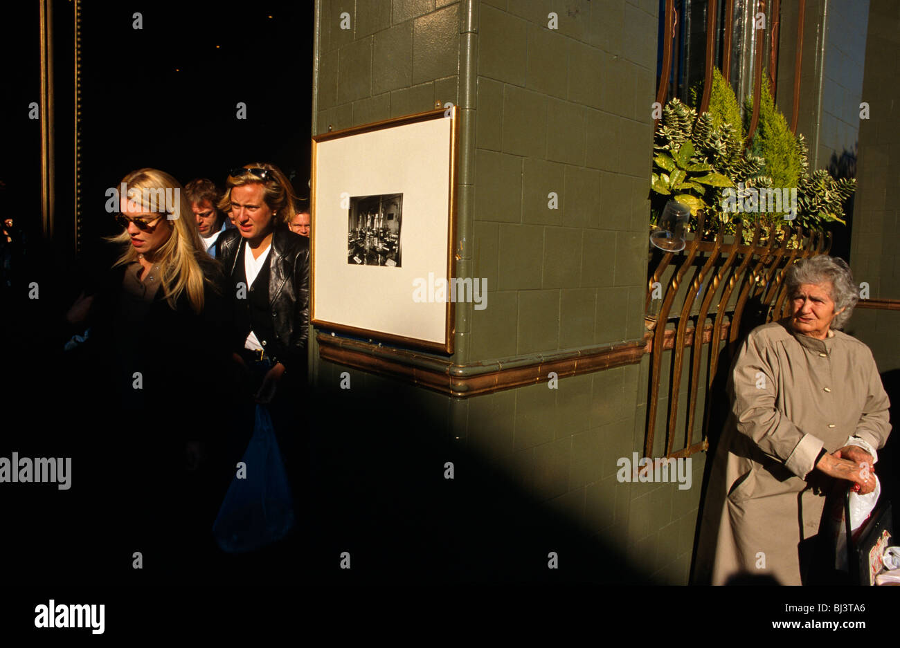 Good-looking trendy young people spill out of a pub as a lonely-looking old lady watches them, mindful of the generation gap. Stock Photo