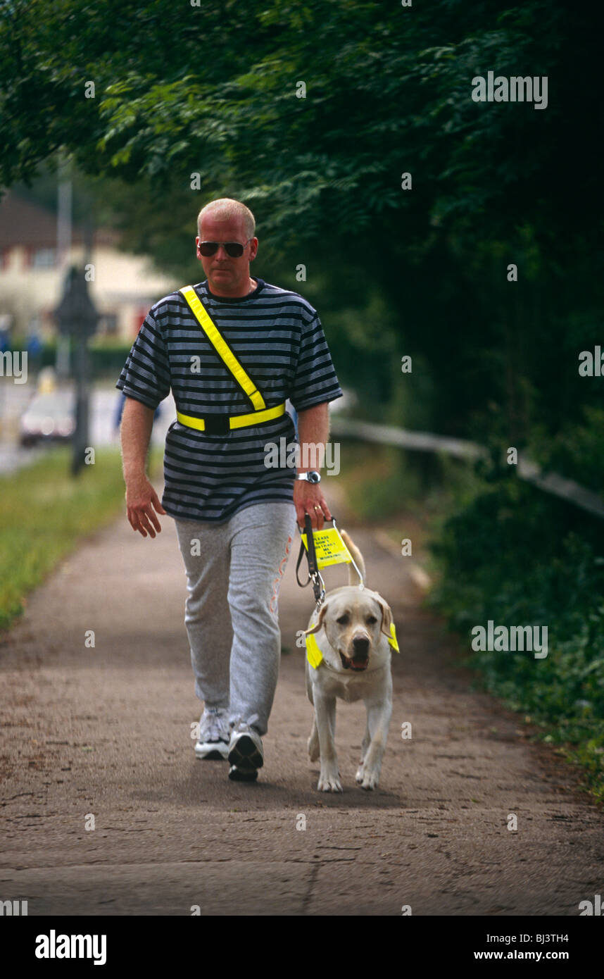 Wearing darkened glasses, an unsighted man walks along a suburban pavement near to the Guide Dogs for the Blind Association. Stock Photo