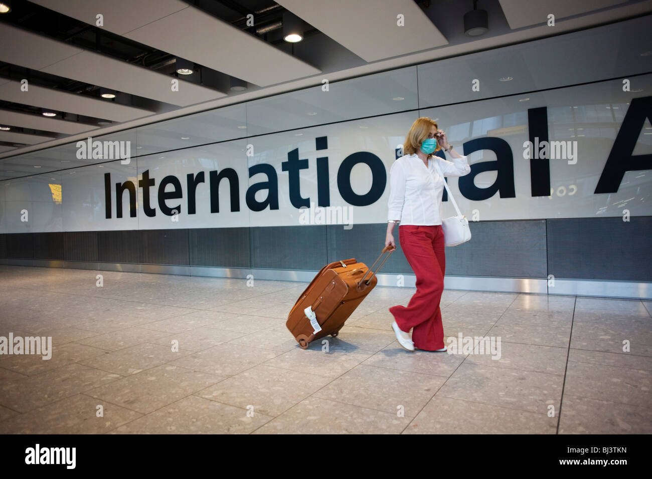 Towing her suitcase after having cleared International Customs, a masked female passenger walks through arrivals at Heathrow T5. Stock Photo