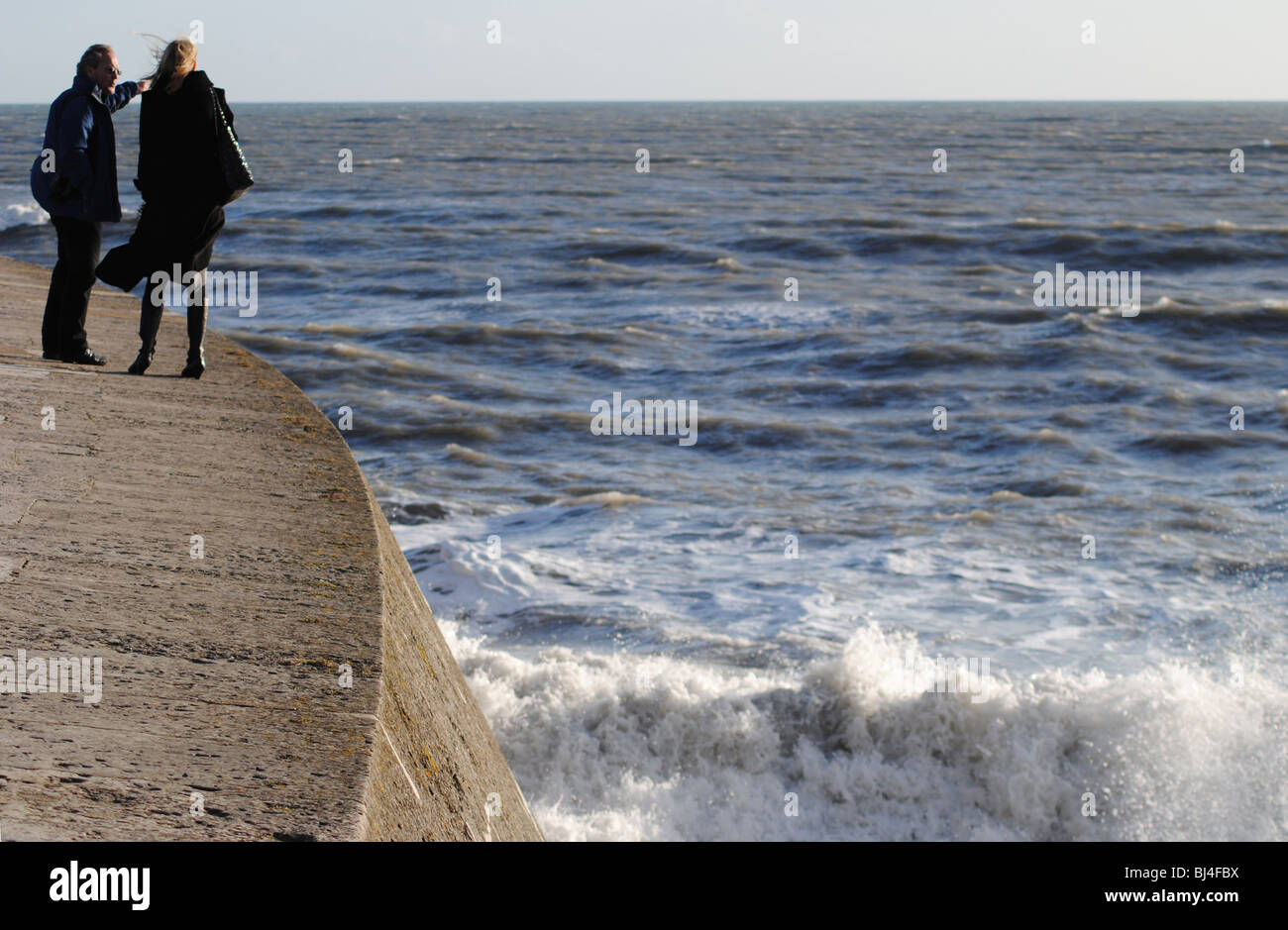 A couple look out over a stormy sea from the Cobb harbor walls, Lyme Regis, Dorset, England Stock Photo