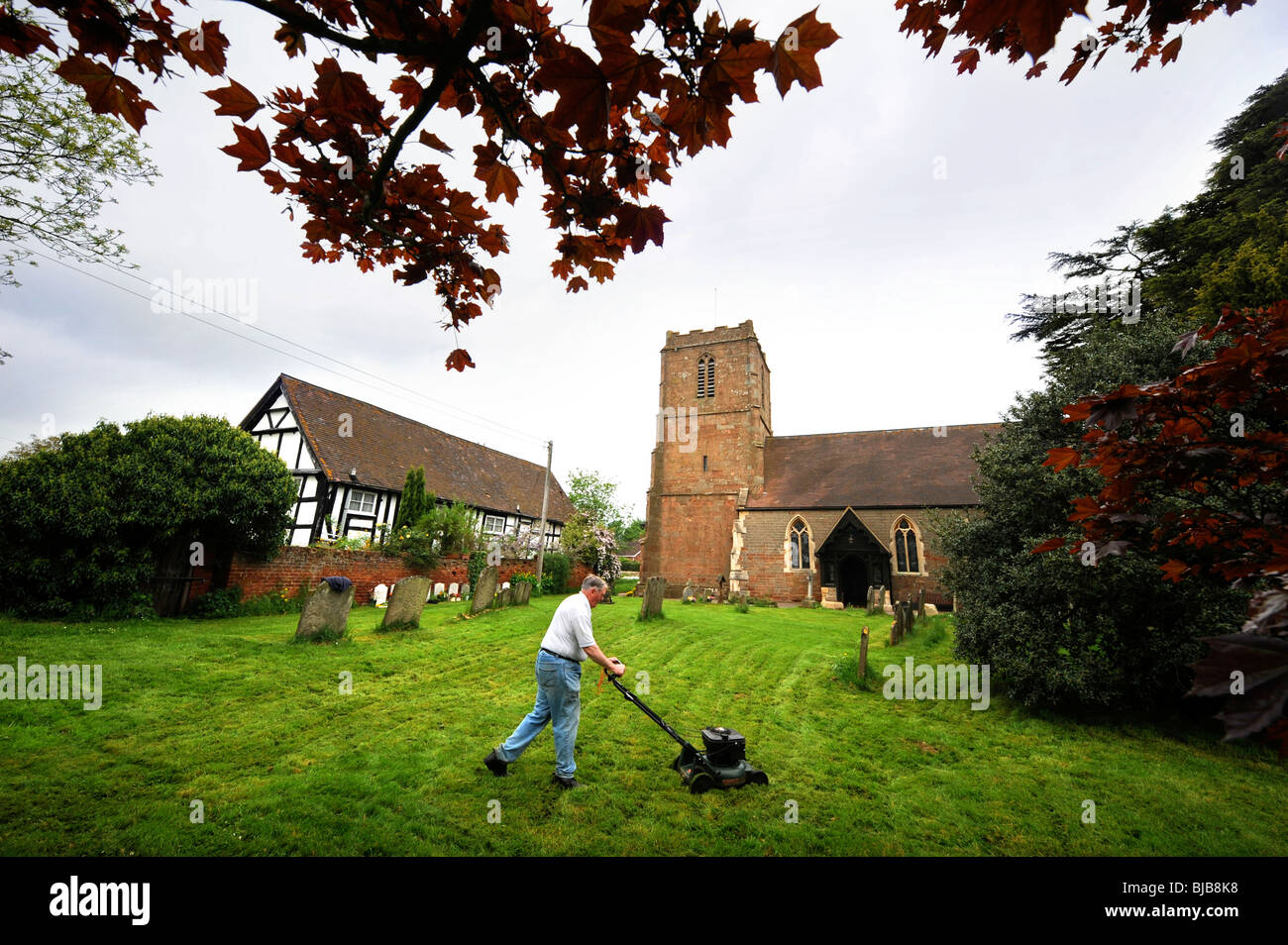 A man mows the grass in the graveyard at The Church of St Bartholomew's in the village of Redmarley D'Abitot, Gloucestershire Stock Photo