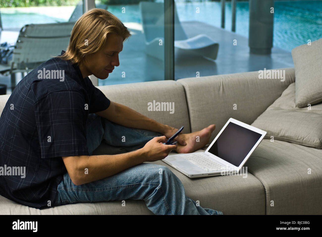 Man sitting on sofa with laptop nearby, using cell phone Stock Photo