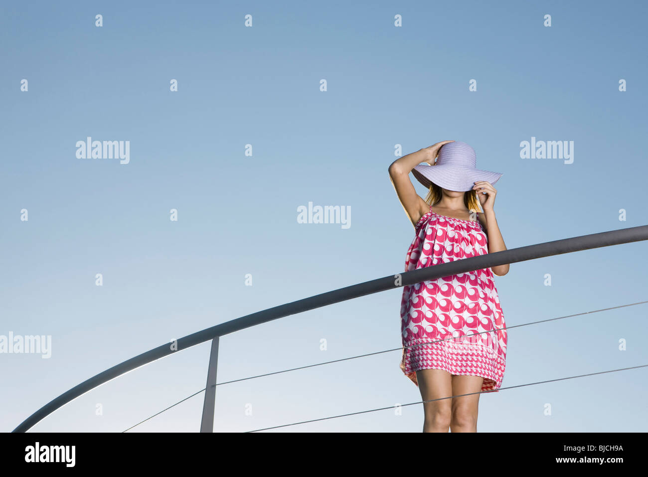 Woman standing on balcony, covering face with sun hat Stock Photo