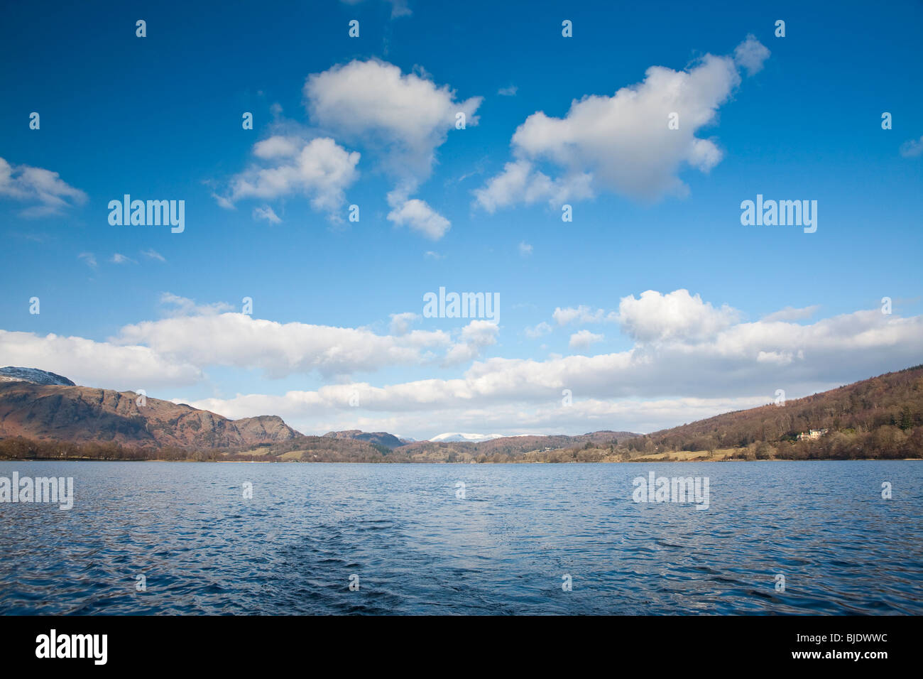 Reflection of sky on Coniston Water in winter with trees and snow covered mountains in background. Stock Photo