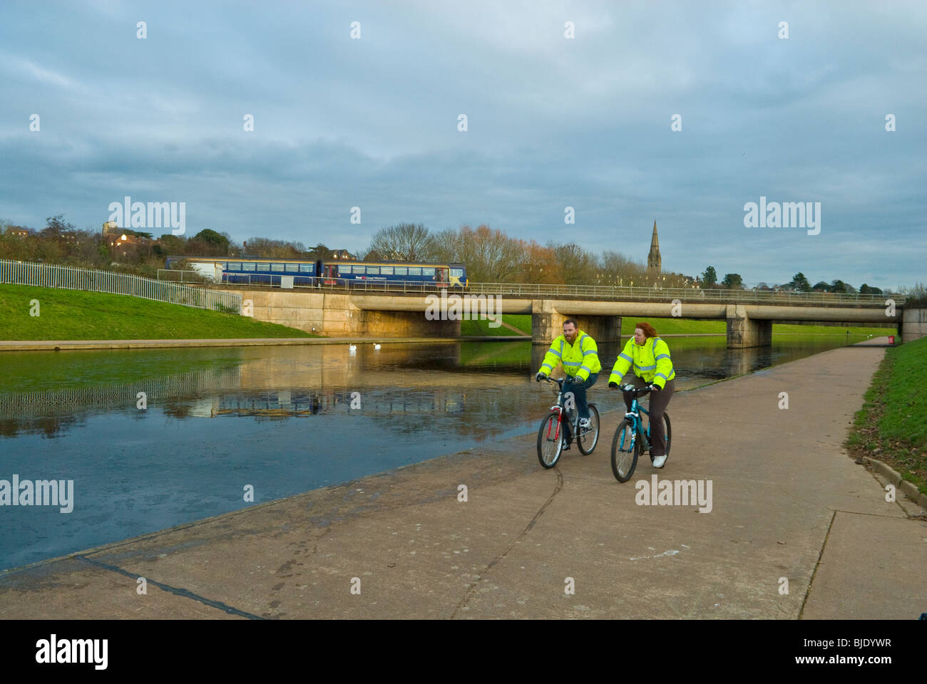 Two cyclists riding along a river path with a train in the background traveling over a bridge in Exeter Devon UK Stock Photo