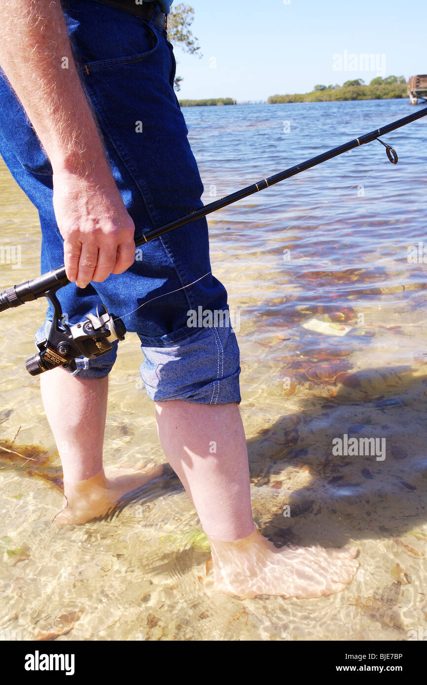 FISHERMAN STANDING IN SHALLOW WATER HOLDING FISHING ROD AND REEL BLUE