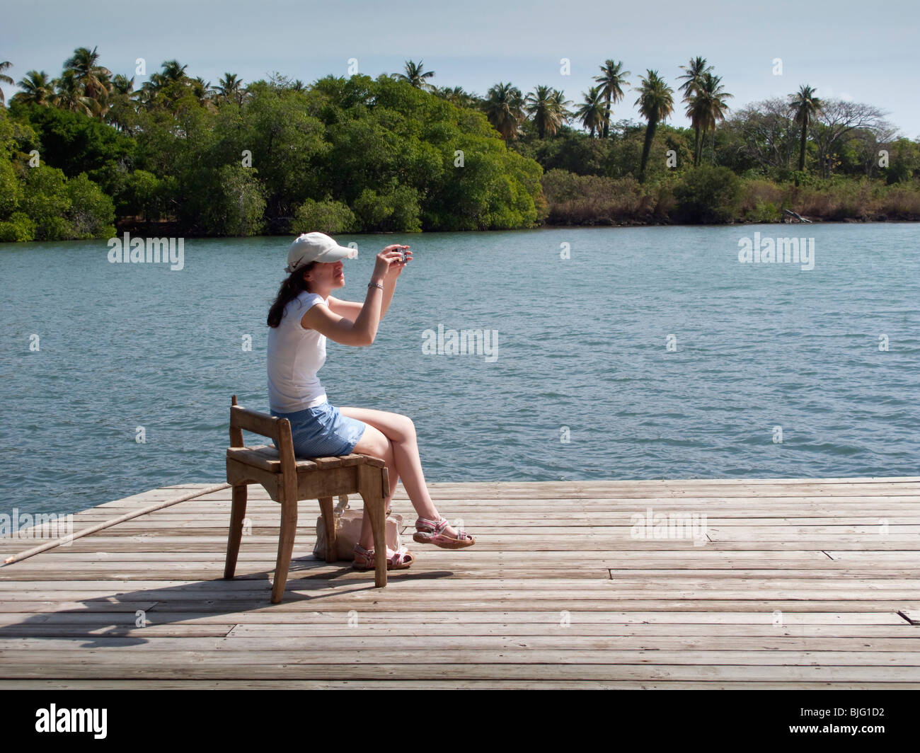 Girl uses old wooden seat on a jetty as support whilst composing a photograph of the Caribbean coastline with palm tree backdrop Stock Photo
