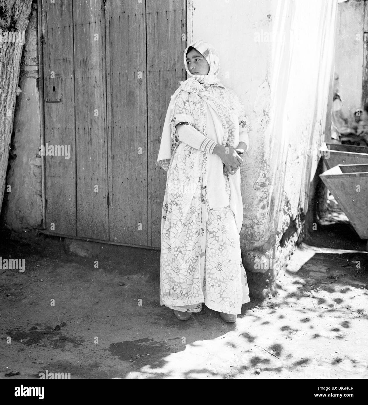 Young lady wearing headdress and long gown standing by old wooden door at corner of stone building, Morocco, 1950s. Stock Photo