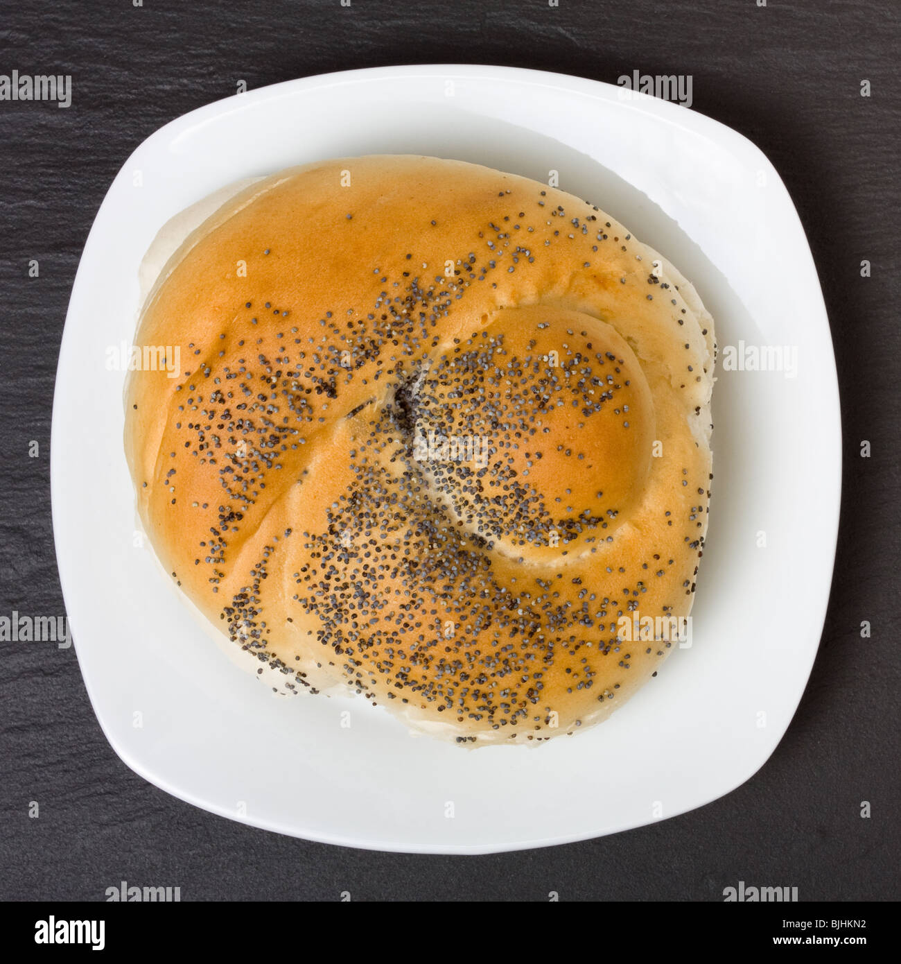 Seeded bread roll on white plate from high viewpoint against dark slate background. Stock Photo