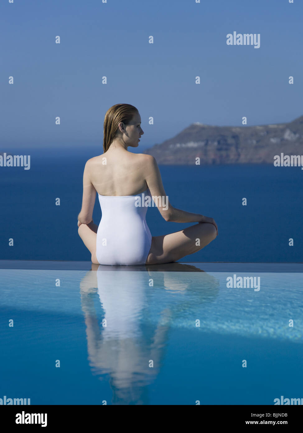 Woman in swimsuit sitting cross legged by infinity pool outdoors Stock Photo