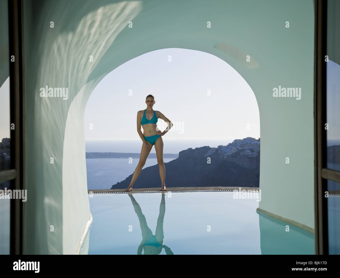 Woman in bikini standing at edge of infinity pool with arch and rock formation Stock Photo