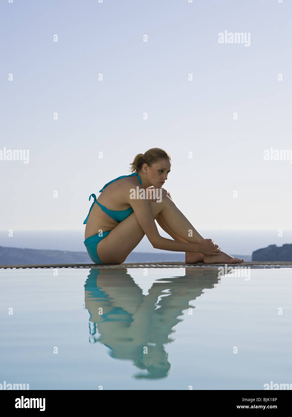 Woman in swimsuit sitting at edge of infinity pool outdoors Stock Photo