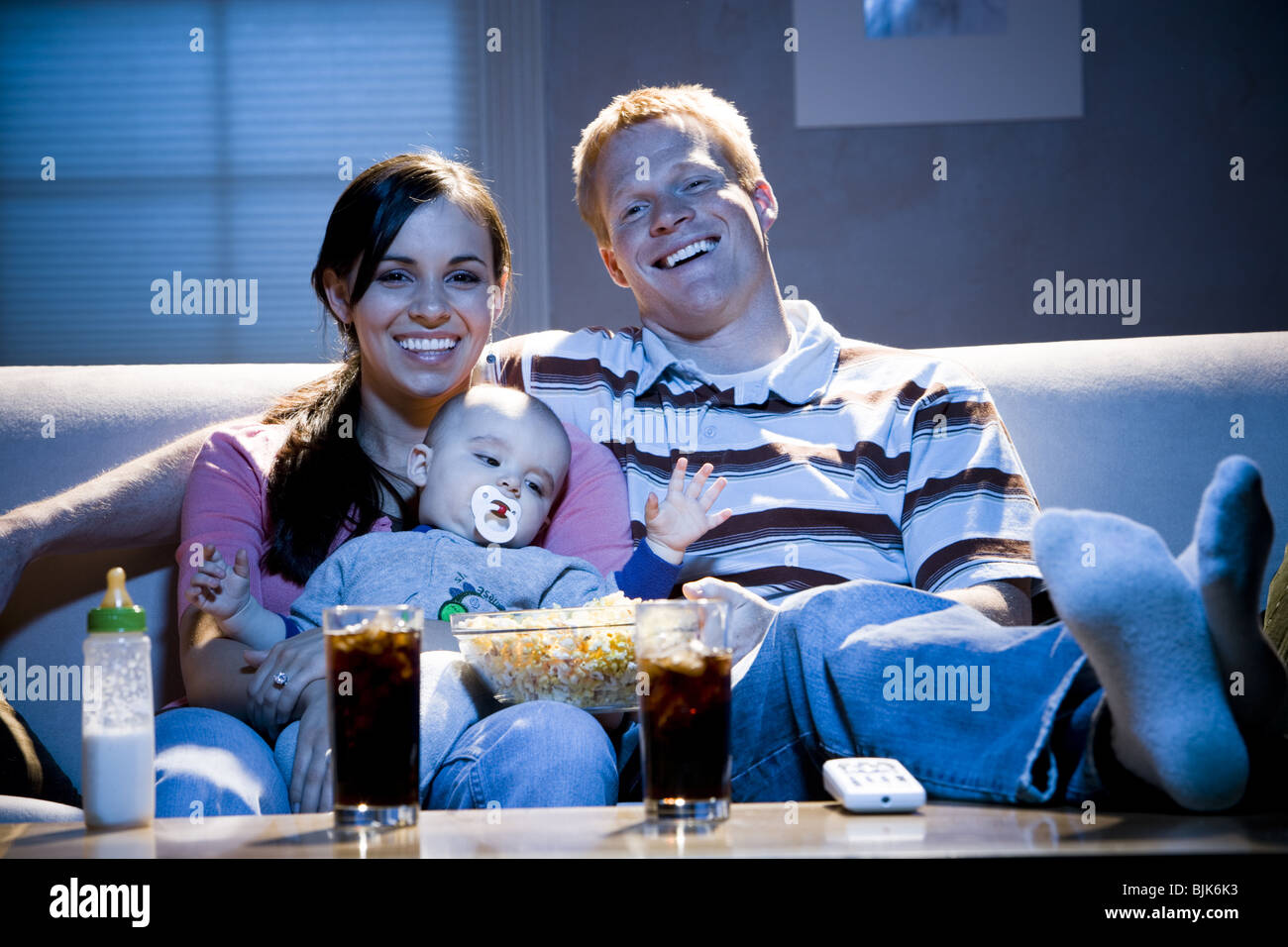 Man on sofa with woman feeding baby and bowl of popcorn smiling Stock Photo