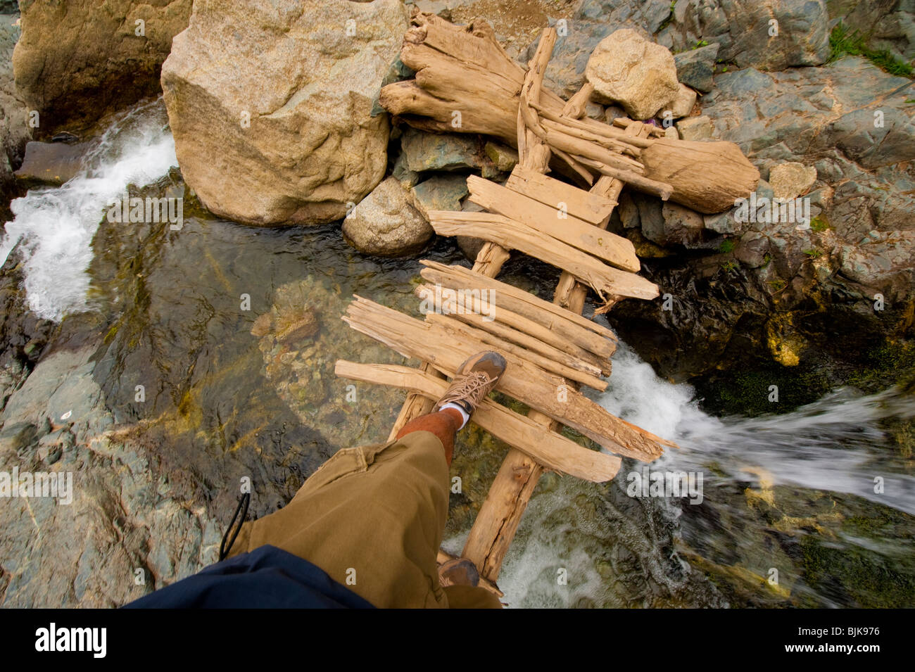 This is an image of a man walking across a wooden bridge over a raging river. Stock Photo