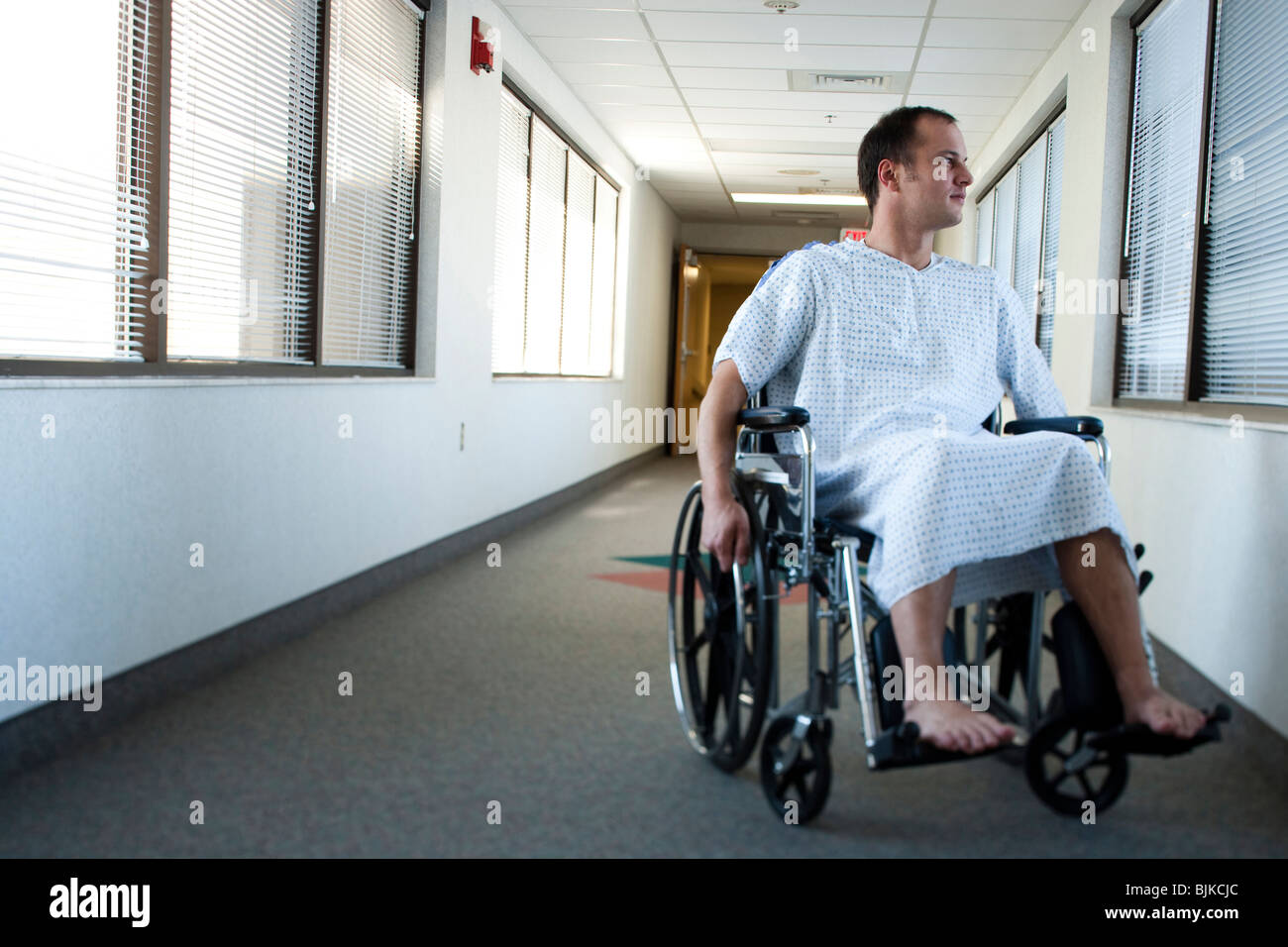 Man in hospital gown in wheelchair in corridor with windows Stock Photo