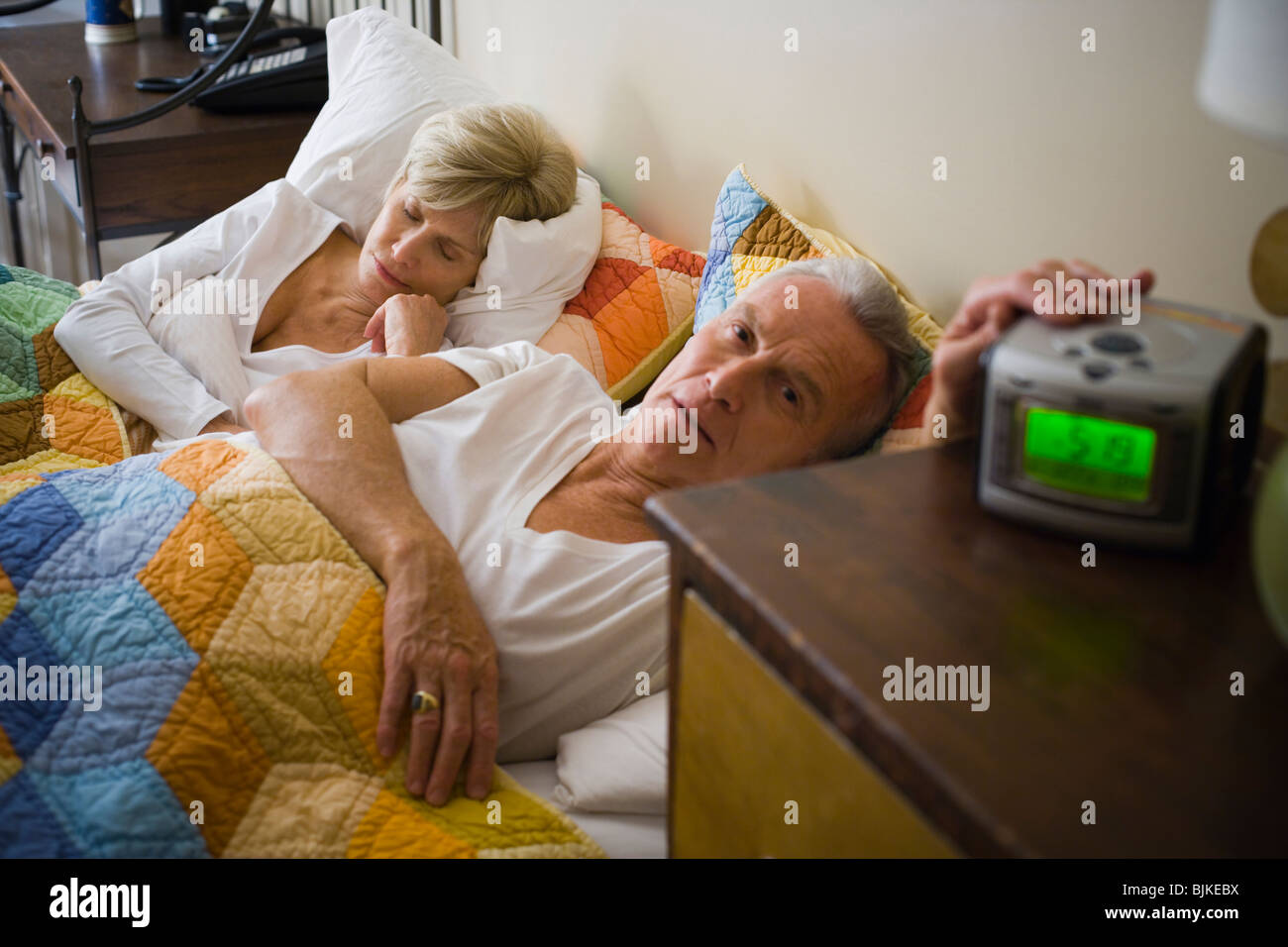 Mature man with alarm clock in bed with sleeping mature woman Stock Photo
