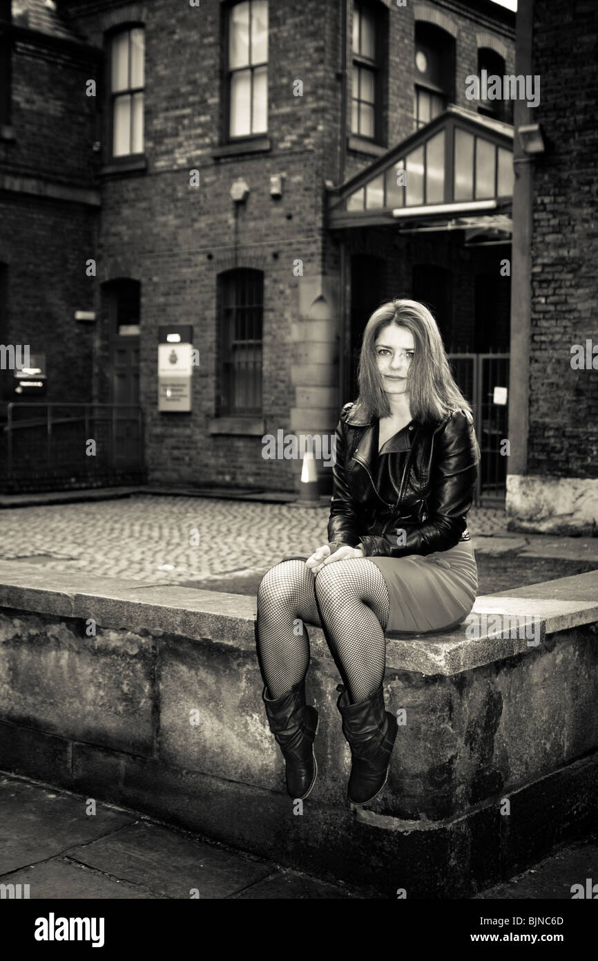 Red head woman in a short skirt and leather jacket and long hair sat on a wall around a courtyard stares at the photographer Stock Photo