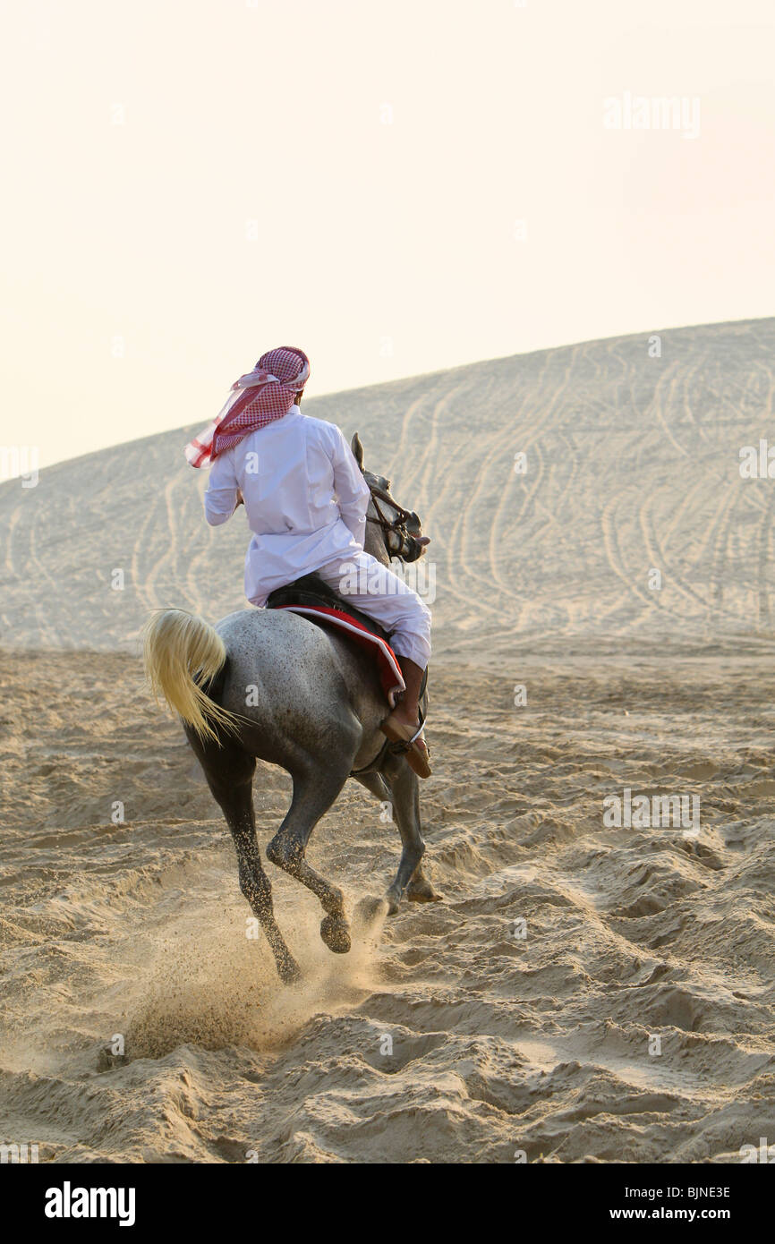 An anonymous Arab man in traditional clothing riding his horse in the sand of a desert bathed in golden sunlight Stock Photo