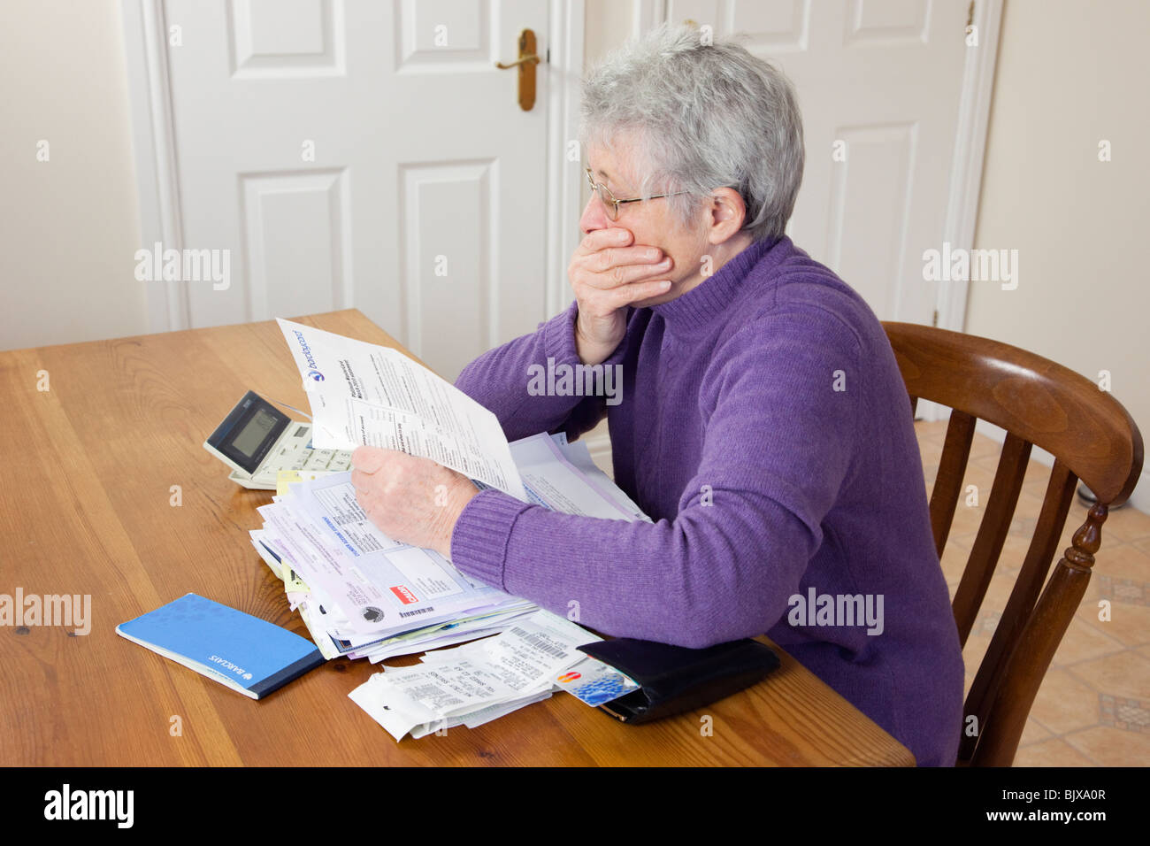 Worried senior woman pensioner with a big pile of bills with hand over her mouth looking shocked at a large credit card bill. England UK Stock Photo